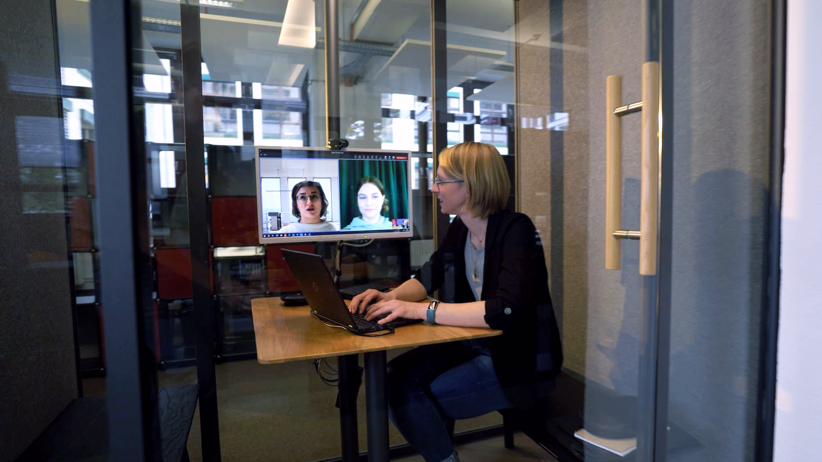 Foto von Rebecca Maier, IT-Administratorin bei der Bundesdruckerei GmbH, die im Büro in einer Meetingzelle sitzt und eine Videokonferenz mit Kollegen und Kolleginnen führt