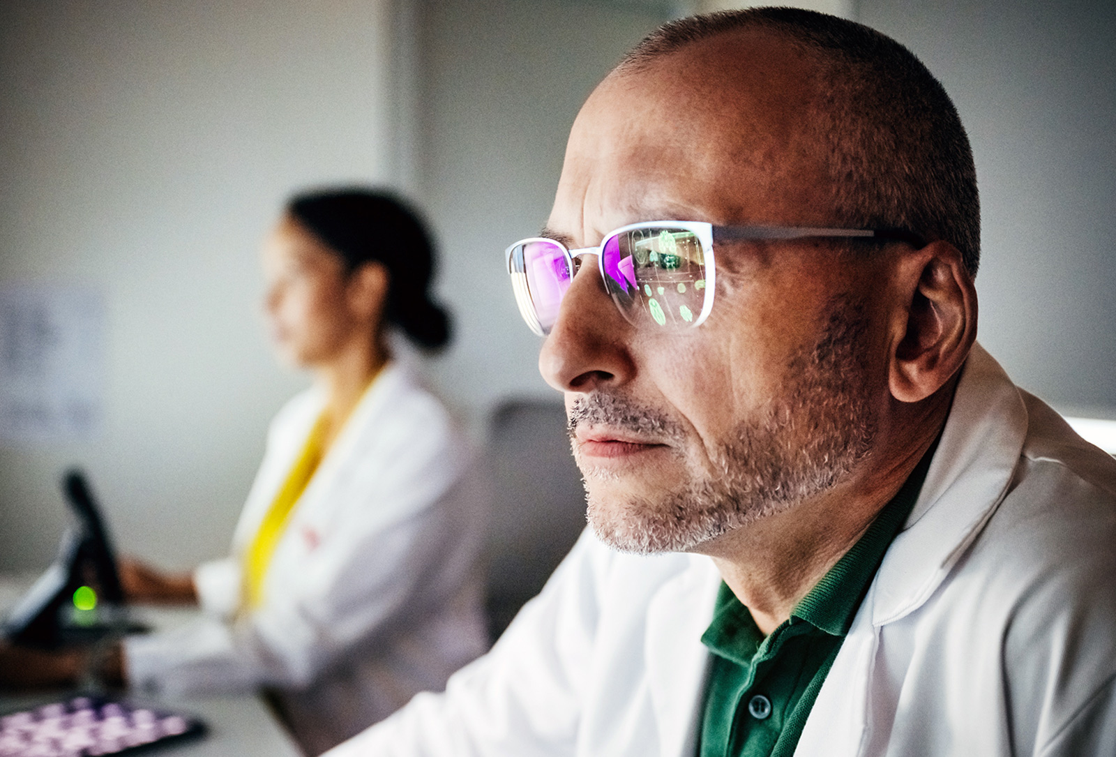  A man with glasses wearing a white coat while sitting at work.