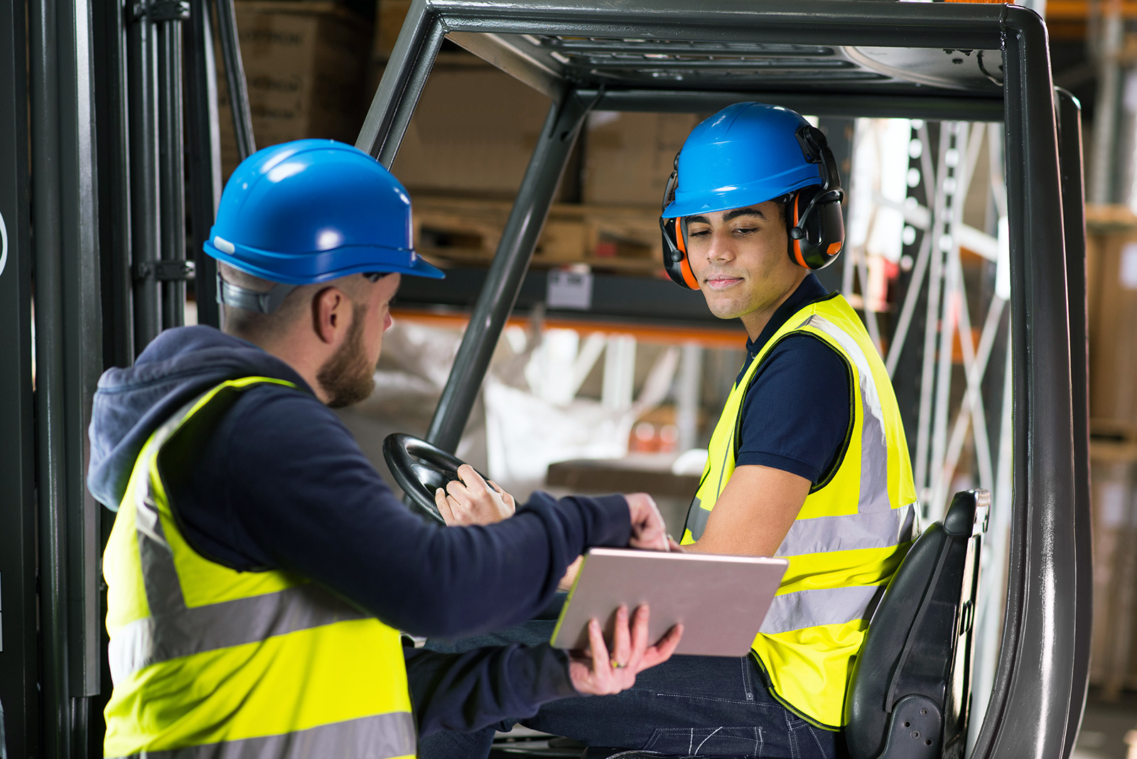 Two people are working together in the warehouse, wearing yellow high-visibility vests and blue helmets.