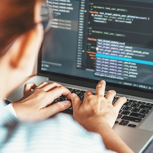 Photo of a person typing on a laptop. Programming code is visible on the screen. 