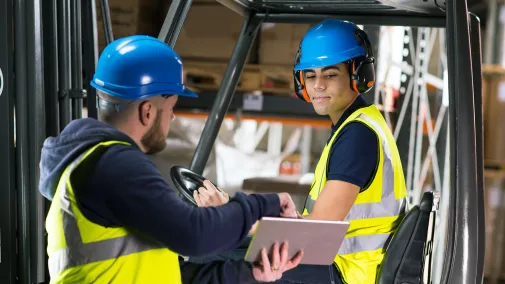 Zwei Personen führen gemeinsam Lagertätigkeiten aus und tragen dabei gelbe Warnwesten sowie blaue Helme.