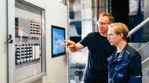 Two employees are standing in front of a system and looking at a screen.