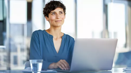 Foto einer Frau mittleren Alters mit kurzen braunen Haaren und blauer Bluse, die an einem Schreibtisch vor einem Laptop sitzt und in Gedanken in die Ferne blickt