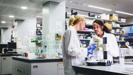 Foto von zwei Forscherinnen in einem Labor, die vor einem Mikroskop stehen und sich unterhalten