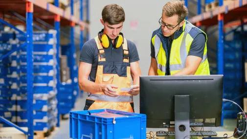 A trainee in warehouse logistics checks stock levels in the warehouse while an experienced employee instructs and supports him.