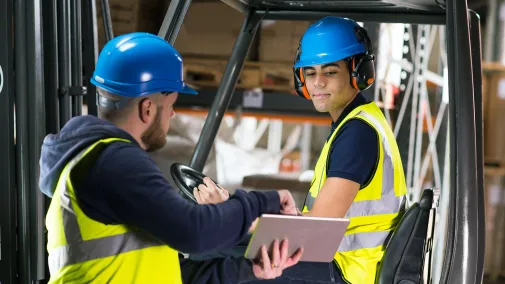 Two people are working together in the warehouse, wearing yellow high-visibility vests and blue helmets.