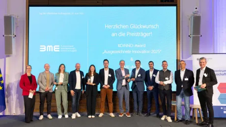 Foto bildet Gewinner des KOINNO ab. Die Personengruppe steht vor einem großen blauen Screen mit Herzlichen Glückwunsch