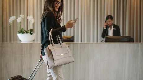 Foto einer Frau, die an einer Hotelrezeption mit einen Rollkoffer vorbeigeht und dabei auf ihr Smartphone blickt