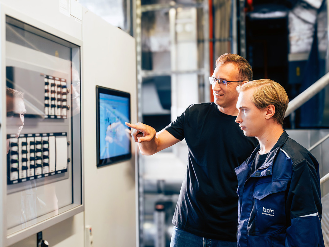 Two employees are standing in front of a system and looking at a screen.