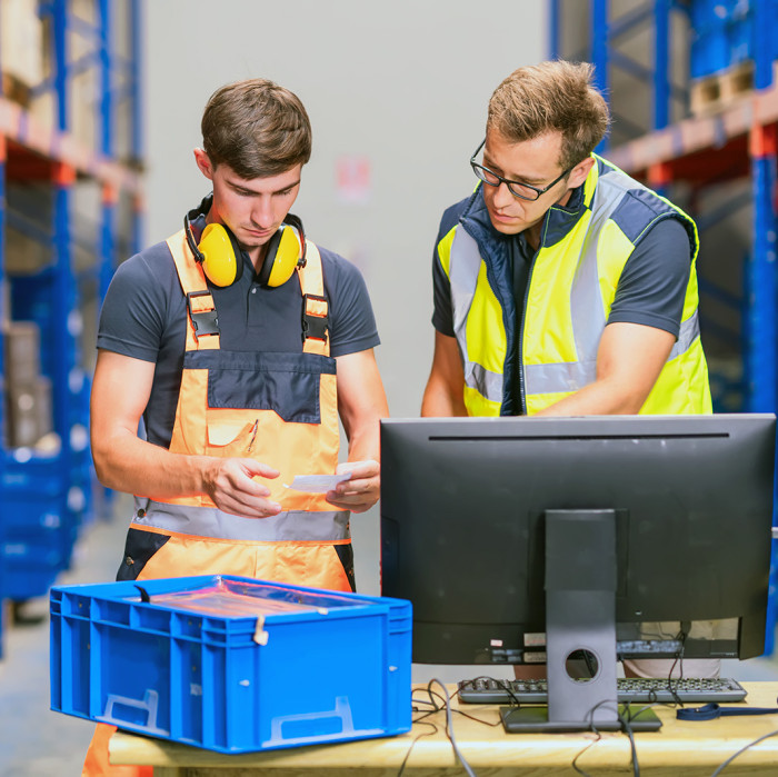 A trainee in warehouse logistics checks stock levels in the warehouse while an experienced employee instructs and supports him.