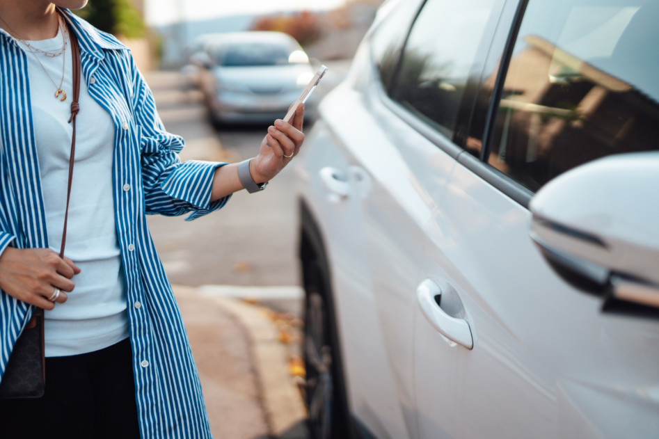 Eine Frau steht vor ihrem Auto und hält ihr Smartphone in der Hand.